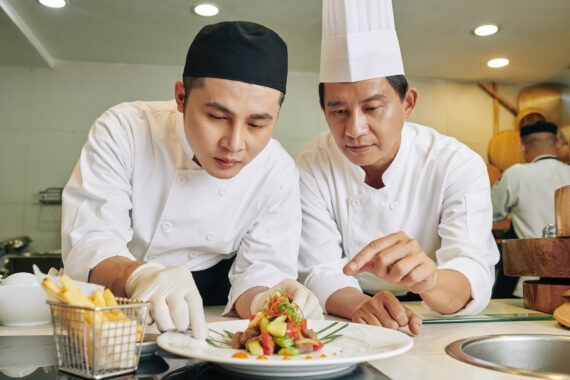 Asian mature chef holding master class for his student he teaching him to decorate the dish in the kitchen